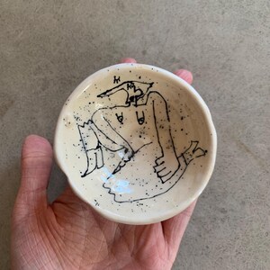 Könnte beinhalten: Eine kleine, runde, cremeweiße Keramikschale mit einer schwarzen Tuschezeichnung einer sitzenden Figur mit spitzem Kopf. Die Oberfläche der Schale ist mit verstreuten schwarzen Sprenkeln versehen, die ein strukturiertes Aussehen im Inneren erzeugen.