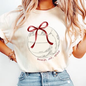 Può includere: T-shirt color crema con un disegno di pallavolo con un fiocco rosso e le parole "bump, set, spike" in un font script. La maglietta è abbinata a jeans di denim blu chiaro.