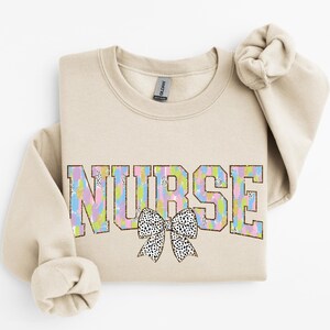 Könnte beinhalten: Ein beiger Pullover mit dem Wort "NURSE" in bunten Buchstaben mit einer schwarz-weißen gepunkteten Schleife. Die Buchstaben sind in Gold umrandet.