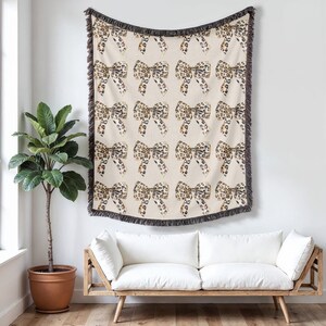 Puede incluir: Una manta de tiro con un fondo beige y un patrón repetido de lazos de estampado de leopardo. La manta tiene un borde de flecos marrones.