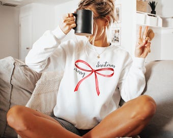 Könnte beinhalten: Weißer Pullover mit einer roten Schleife und dem Aufdruck "merry christmas" auf der Vorderseite.
