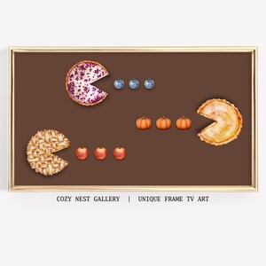 Puede incluir: Una impresión enmarcada con un diseño inspirado en Pac-Man, utilizando rebanadas de pastel como personajes de Pac-Man. Los "Pac-Man" están hechos de rebanadas de pastel, y los "puntos" son arándanos, manzanas y calabazas. El texto "COZY NEST GALLERY | UNIQUE FRAME TV ART" está en la parte inferior.