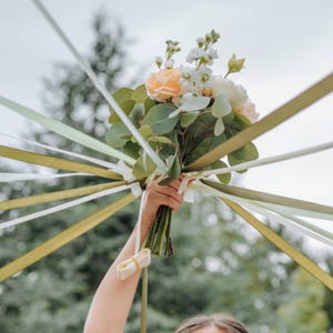 Peut inclure: Un bouquet de fleurs avec des roses pêche et blanches, des giroflées blanches et du feuillage vert. De longs rubans verts, blancs et dorés s'étendent du bouquet. L'arrière-plan flou montre des arbres verts et un ciel nuageux, pour un effet festif.