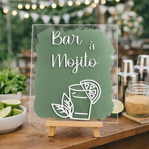 Peut inclure: Panneau en acrylique transparent avec l'inscription "Bar à Mojito" en écriture blanche. Le panneau présente un fond vert et un contour blanc d'un cocktail mojito. Le panneau est présenté sur un petit chevalet en bois.