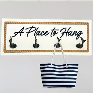 May include: A Place to Hang wall decor with black hooks, a striped tote bag, and navy blue pillows on a wooden bench. A white fringed throw is draped on the bench. The wall decor has a rope border.