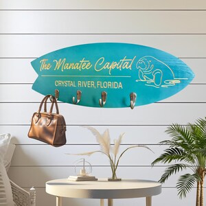 May include: A turquoise surfboard-shaped wall hook with the text "The Manatee Capital Crystal River, Florida" and a manatee illustration. A brown leather handbag hangs from one of the hooks. The wall is white with horizontal wood paneling.