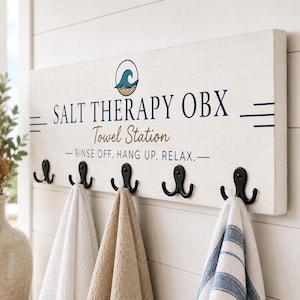 May include: A rectangular white wooden sign with five black metal double hooks. The sign features a blue wave graphic, the text SALT THERAPY OBX, Towel Station, and RINSE OFF. HANG UP. RELAX. Three towels in white, tan, and blue-striped patterns hang below.