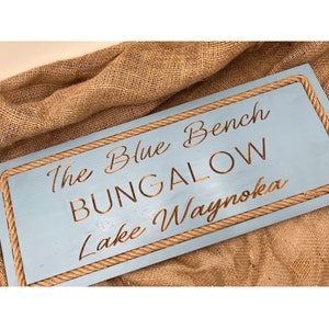 May include: A rectangular, light blue wooden sign with the words "The Blue Bench BUNGALOW Lake Waynoka" in a brown, engraved font. The sign is framed with a rope border and rests on a burlap surface.
