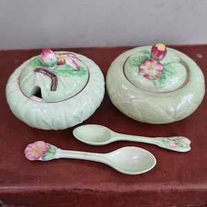 May include: Two light green ceramic lidded jars and two spoons. The lids are decorated with pink and green floral designs. One jar has a spoon slot. The spoons have floral accents on the handles. The set is on a brown surface.