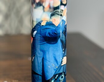 Puede incluir: Un vaso de acero inoxidable con fondo rojo y una foto de dos hombres abrazándose. La foto muestra a un hombre con un traje azul con el brazo alrededor de otro hombre que lleva una camisa negra. El fondo de la foto es una multitud borrosa de personas.