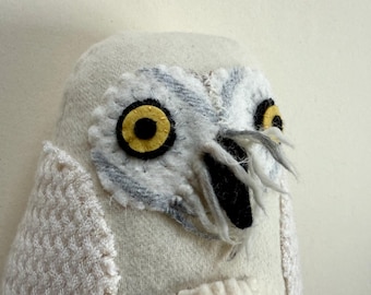 May include: A handmade plush owl with a cream-colored body and detailed facial features. The owl's eyes are large, with yellow irises and black pupils. The face has gray and white accents, and the beak is black. The wings are textured.
