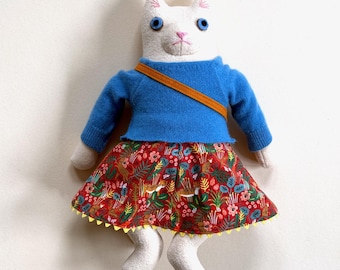 May include: A stuffed cat doll with white fabric, blue button eyes, and pink stitched nose and mouth. It wears a long-sleeved blue sweater with an orange crossbody strap and a red skirt featuring a floral and tiger print with a yellow scalloped hem.