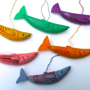May include: Six felt fish ornaments in various colors, including orange, yellow, green, pink, purple, and blue. Each fish has a different embroidered design, including a skeleton, scales, and a grid pattern. The fish are hanging from strings.