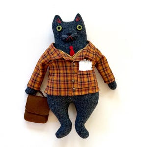 May include: A handmade plush cat doll with a gray body, wearing a plaid shirt in shades of orange, brown, and black, a red tie, and carrying a brown satchel. The cat has green eyes and red embroidered details.
