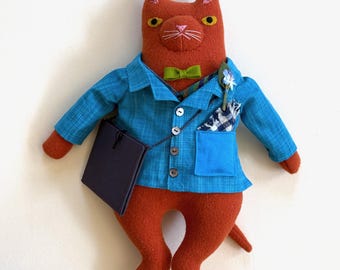 May include: A plush orange cat doll wearing a blue button-down shirt with a subtle grid pattern, a green bow tie, and a dark purple shoulder bag. The cat has embroidered facial features, a small blue flower in its pocket, and a pocket square.