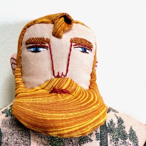 May include: A handmade fabric doll head with embroidered features. The doll has blue eyes, a red nose and mouth, and a large orange and yellow striped beard. The doll's hair is also orange and yellow. The doll is wearing a patterned shirt.