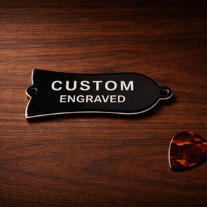 May include: A black, custom engraved guitar pick holder with the text "CUSTOM ENGRAVED" in white lettering. A tortoiseshell guitar pick is next to the holder. The items are on a dark wood surface.