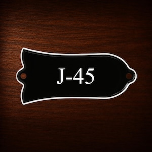 May include: A black bell-shaped guitar truss rod cover with a white border and the white text J-45 printed in the center. The cover features two small circular mounting holes on the left and right sides for attachment to a guitar headstock.