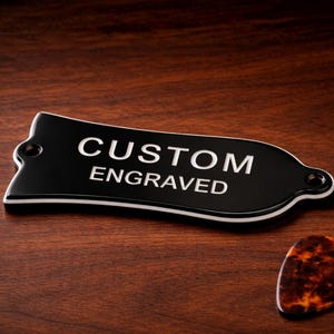 May include: A black guitar headstock plate with the words "CUSTOM ENGRAVED" in white lettering. The plate has a white border and two holes for mounting. A tortoiseshell guitar pick rests nearby on a dark wood surface.