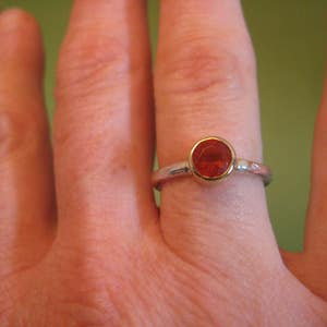 Puede incluir: Un anillo de plata con una piedra preciosa roja engastada en un bisel de oro. El anillo se lleva en un dedo.