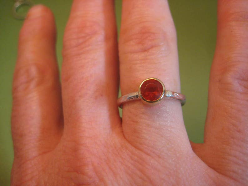Puede incluir: Un anillo de plata con una piedra preciosa roja engastada en un bisel de oro. El anillo se lleva en un dedo.