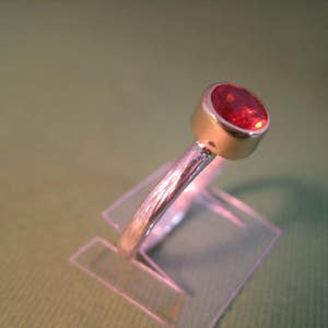 Puede incluir: Un anillo de plata con una piedra preciosa roja engastada en un engaste de bisel. El anillo se muestra en un soporte de pl&aacute;stico transparente.