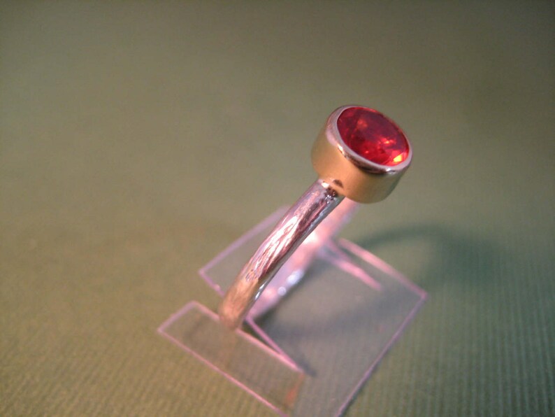 Puede incluir: Un anillo de plata con una piedra preciosa roja engastada en un engaste de bisel. El anillo se muestra en un soporte de pl&aacute;stico transparente.