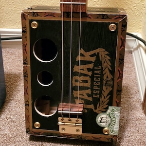 May include: A handmade cigar box guitar with a dark green front featuring the text "TABAK ESPECIAL" and a gold laurel wreath design. The guitar has a wooden neck, strings, and a gold-colored bridge.