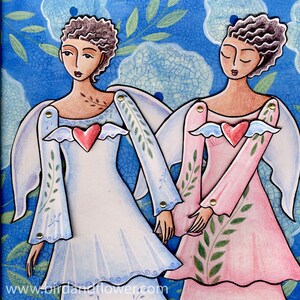 May include: Two paper angels with wings, one in blue and one in pink, with a heart on their chest. The angels have brown hair and are holding hands.