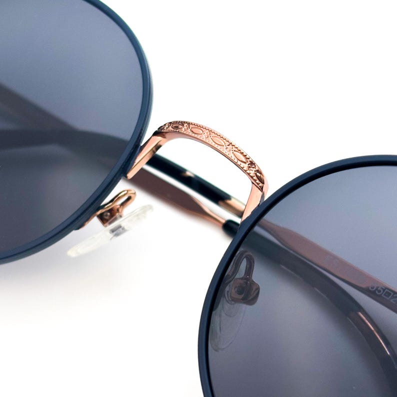 Pu&ograve; includere: Primo piano della montatura in metallo color oro rosa di un paio di occhiali da sole rotondi con lenti blu scuro. La montatura presenta un design decorativo.