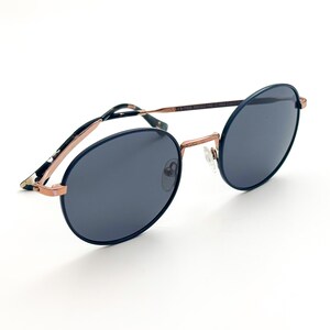 Pu&ograve; includere: Occhiali da sole rotondi blu con montatura in metallo color oro rosa. Gli occhiali hanno lenti scure.