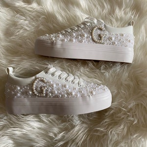 Peut inclure: Baskets blanches à plateforme ornées de fausses perles. Chaque chaussure présente un "G" incrusté de perles sur le côté. Les baskets ont des lacets blancs et sont posées sur un fond blanc duveteux.