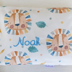 Pode incluir: Uma fronha retangular branca com um padrão repetitivo de rostos de leão de desenho animado com jubas multicoloridas, folhas verde-azuladas e pequenos pontos. O nome Noah está bordado em fio azul sobre o tecido.
