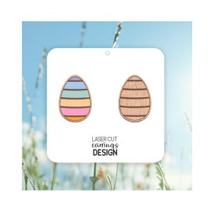 May include: A pair of laser cut wooden earrings shaped like Easter eggs. One earring is painted with pastel stripes and the other is a natural wood colour. The earrings are displayed on a white card with the text "LASER CUT earrings DESIGN".