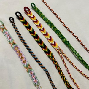 May include: Six colourful friendship bracelets with different patterns, including chevron, diamond, and striped designs. The bracelets are made with embroidery floss and have tassels at the ends.