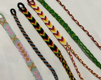 Könnte beinhalten: Sechs handgefertigte, gewebte Freundschaftsarmbänder mit verschiedenen Mustern und Farben. Ein Armband hat ein pastellfarbenes Rautenmuster, ein anderes hat schwarze und blaue diagonale Streifen, und weitere zeigen Zickzackmuster in Rot, Gelb, Grün und Weiß.