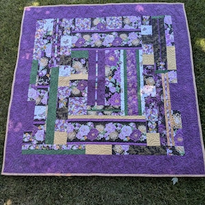 Könnte beinhalten: Eine handgefertigte Steppdecke mit einem lila Rand und einem zentralen Design mit floralen Mustern in Lila-, Grün- und Goldtönen. Die Steppdecke liegt auf einer grasbewachsenen Oberfläche und zeigt ihre komplizierten Patchwork- und Steppdetails.