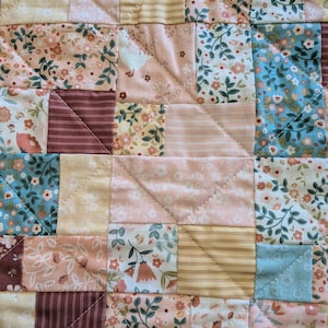 May include: Close-up of a patchwork quilt with a variety of floral and striped patterns in shades of pink, peach, cream, and blue. The quilt is made of squares and triangles, stitched together with a diamond pattern.