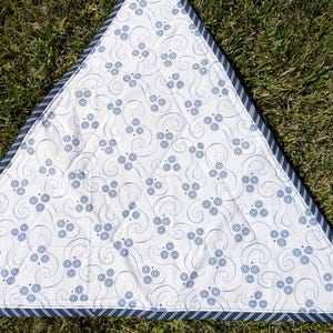 May include: A triangular white and blue patterned quilt. The quilt features a repeating floral design in blue, with a blue and white striped border. The quilt is lying on a bed of green grass.