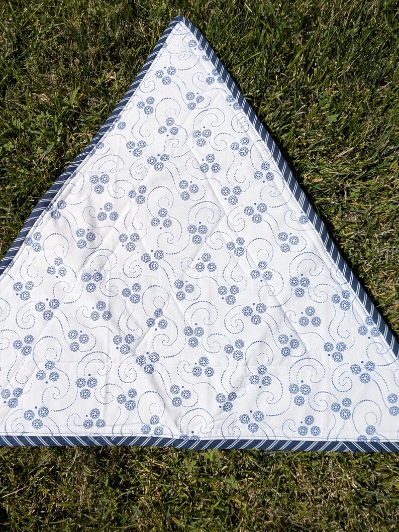 May include: A triangular white and blue patterned quilt. The quilt features a repeating floral design in blue, with a blue and white striped border. The quilt is lying on a bed of green grass.