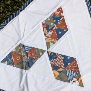 May include: A handmade quilt featuring white fabric with diamond-shaped patchwork in shades of blue, brown, and green. The patchwork has floral and geometric patterns. The quilt is bordered with a blue and white striped fabric.