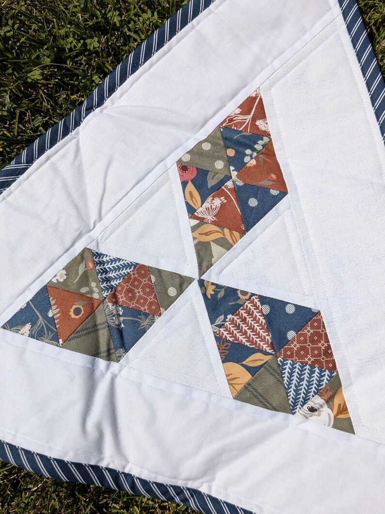 May include: A handmade quilt featuring white fabric with diamond-shaped patchwork in shades of blue, brown, and green. The patchwork has floral and geometric patterns. The quilt is bordered with a blue and white striped fabric.