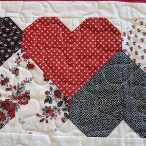 Może przedstawiać: Pikowany bieżnik na stół z dużym czerwonym sercem z białym wzorem w romby. Serce otoczone jest kwadratami patchworkowymi z kwiatowymi i geometrycznymi wzorami w kolorze czerwonym, czarnym i białym. Bieżnik ma czerwoną obwódkę.