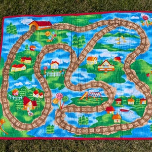 May include: A colorful, quilted play mat featuring a whimsical train track design. The mat showcases a winding brown track through a landscape with houses, trees, and lollipops. The border is red.