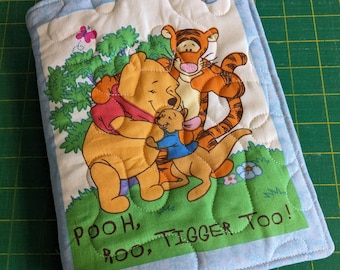 May include: A rectangular quilted fabric cover featuring a printed illustration of Winnie the Pooh, Tigger, and Roo hugging in a green field. The text POOH, ROO, TIGGER TOO! is printed at the bottom. The border is light blue with a subtle pattern.