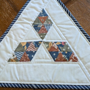 May include: A handmade, triangular quilted placemat. The placemat features a white background with three diamond-shaped sections of colorful, patterned fabric triangles. A navy blue and white striped border frames the edges.
