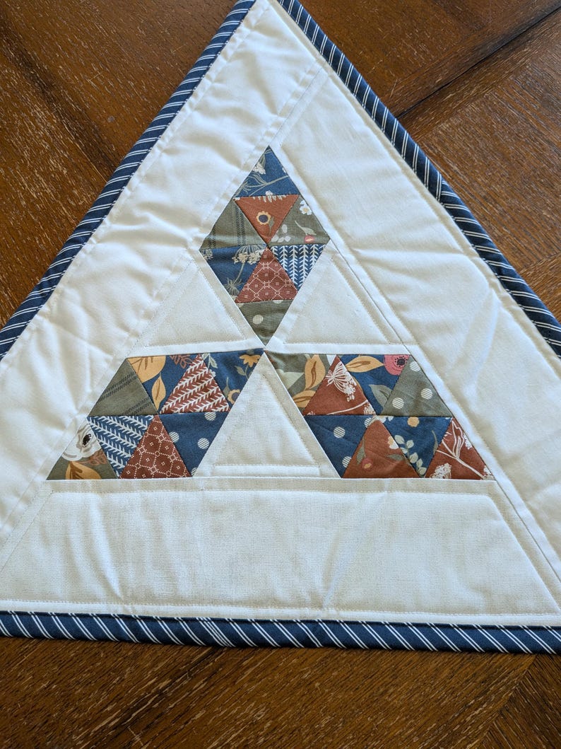 May include: A handmade, triangular quilted placemat. The placemat features a white background with three diamond-shaped sections of colorful, patterned fabric triangles. A navy blue and white striped border frames the edges.