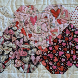 May include: Close-up of a Valentine's Day themed quilt with heart-shaped patterns. The quilt features various fabrics with hearts, love letters, and floral designs in shades of pink, red, and black. The edges are finished with a pink and white striped border.