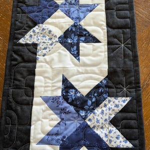 May include: A rectangular quilted table runner featuring a central cream-colored panel with two large star motifs composed of various blue floral and solid fabric patches. The runner is bordered by solid black fabric with stitched starburst patterns.