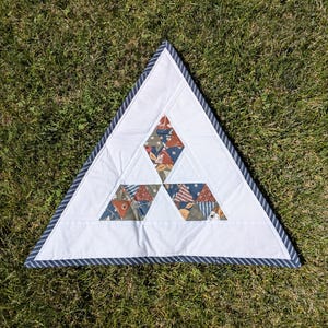 May include: A triangular white quilt with a blue and white striped border. The quilt features three diamond-shaped patches with floral and geometric patterns in shades of orange, blue, and green. The quilt is placed on a bed of green grass.